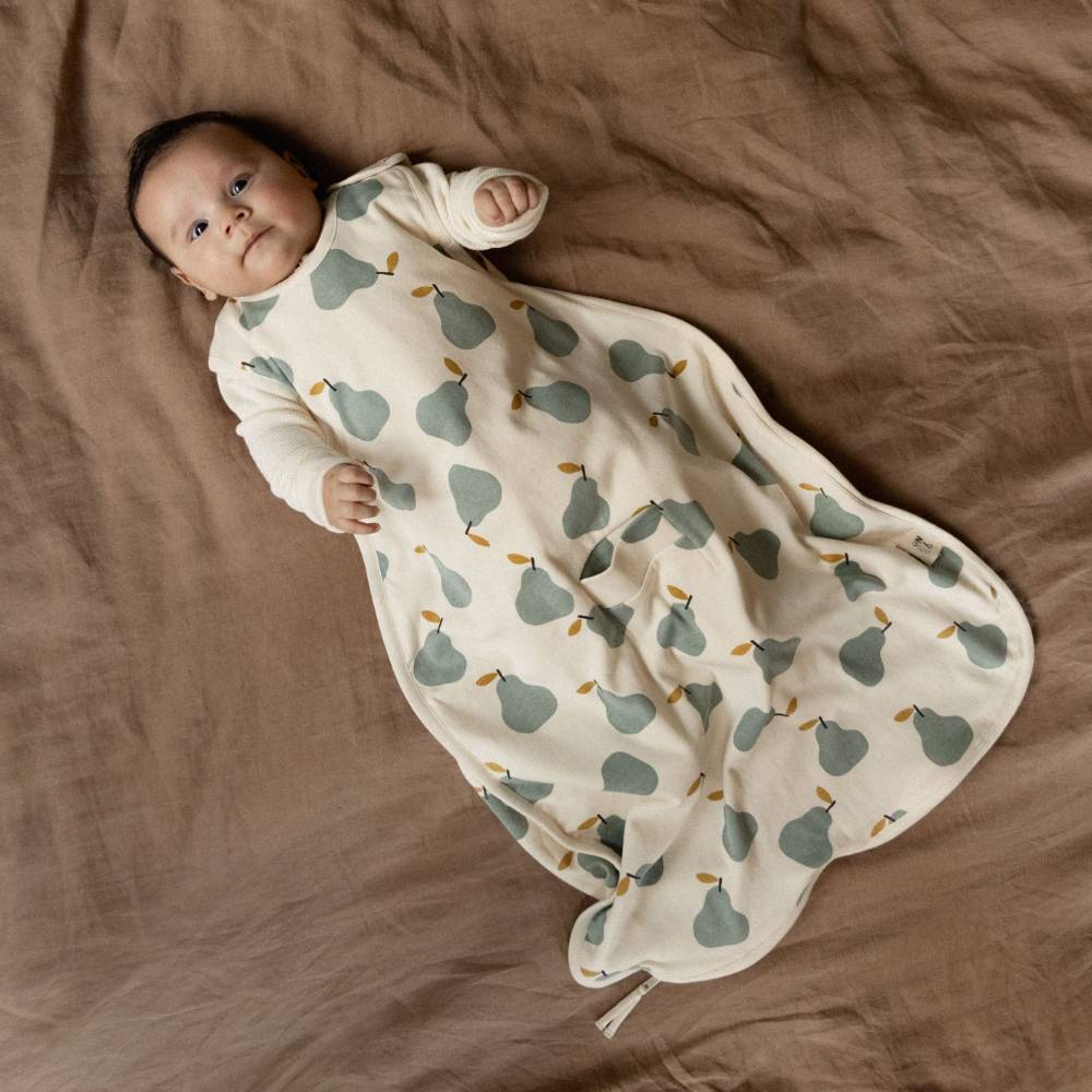 Baby lying on a bed wearing a baby sleeping bag with a pear pattern. 