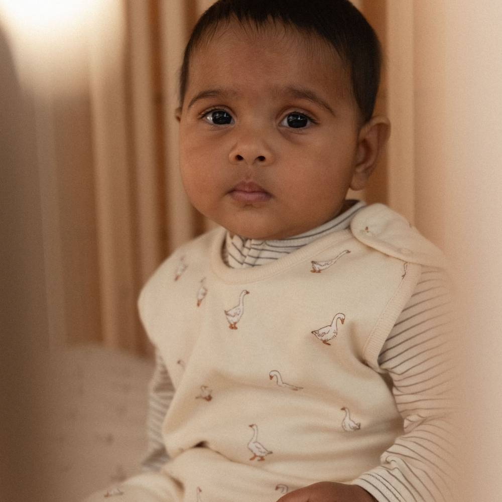 A baby wearing a cream-colored sleep bag with a goose pattern over a striped long-sleeve top, posed against a soft beige background.