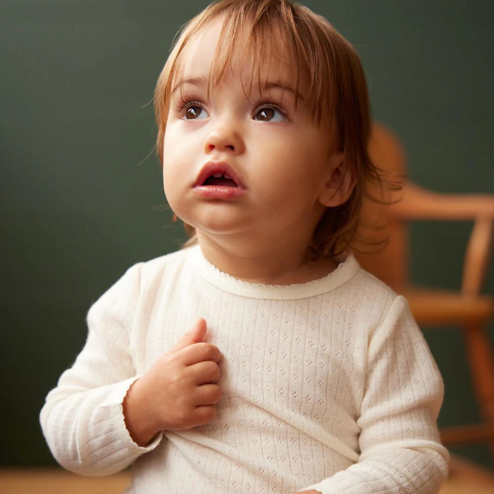 Child wearing a white organic merino cotton pointelle long sleeve sitting on a wooden chair with a forest green background