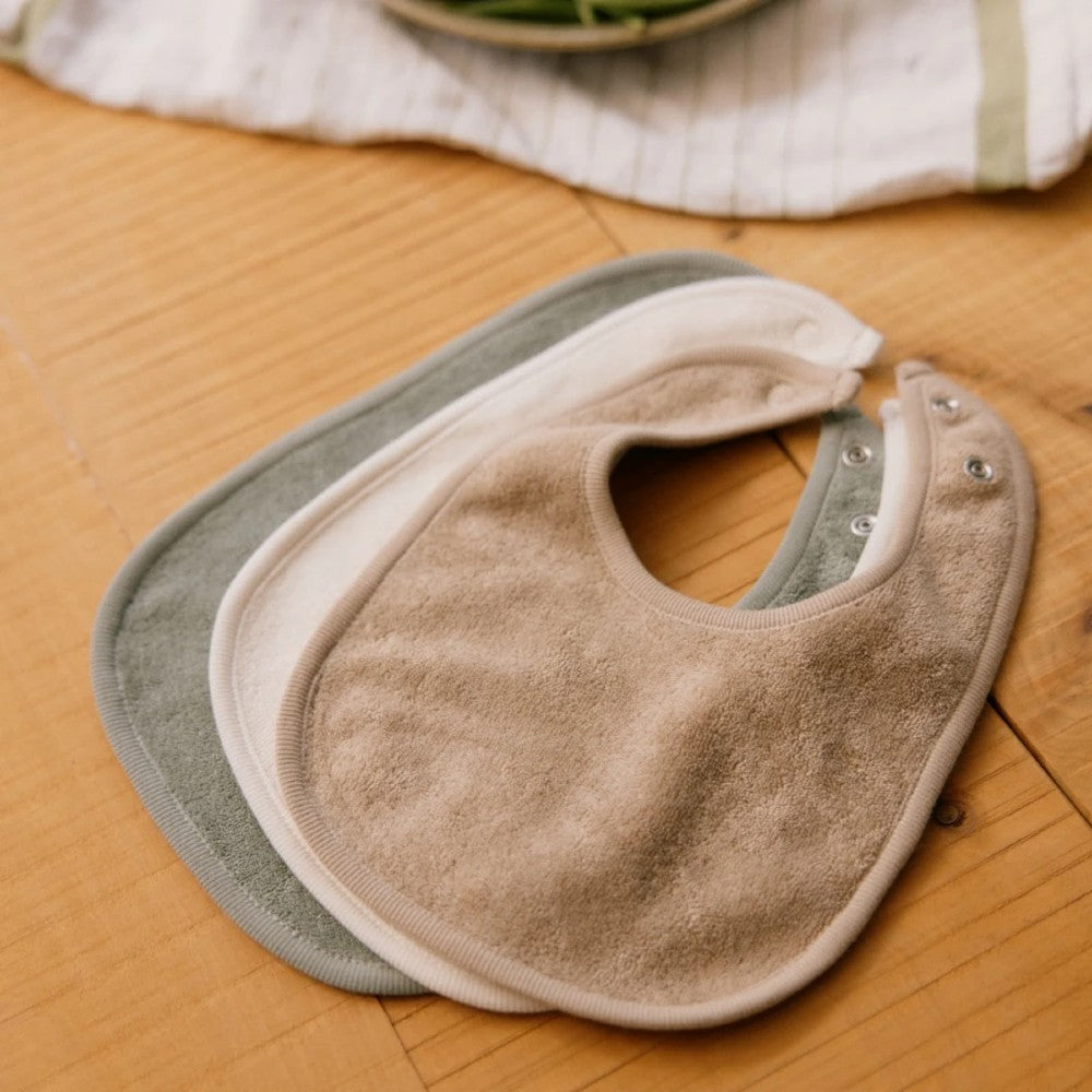Three cotton towelling baby bibs with metal snap domes in beige, cream and green on a wooden surface
