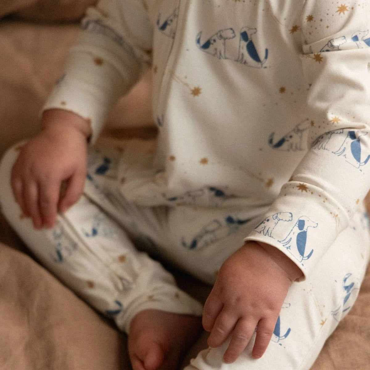 A baby wearing a cream-colored long-sleeve footed sleeper with a blue dog pattern and small gold star accents, sitting on a beige fabric surface.