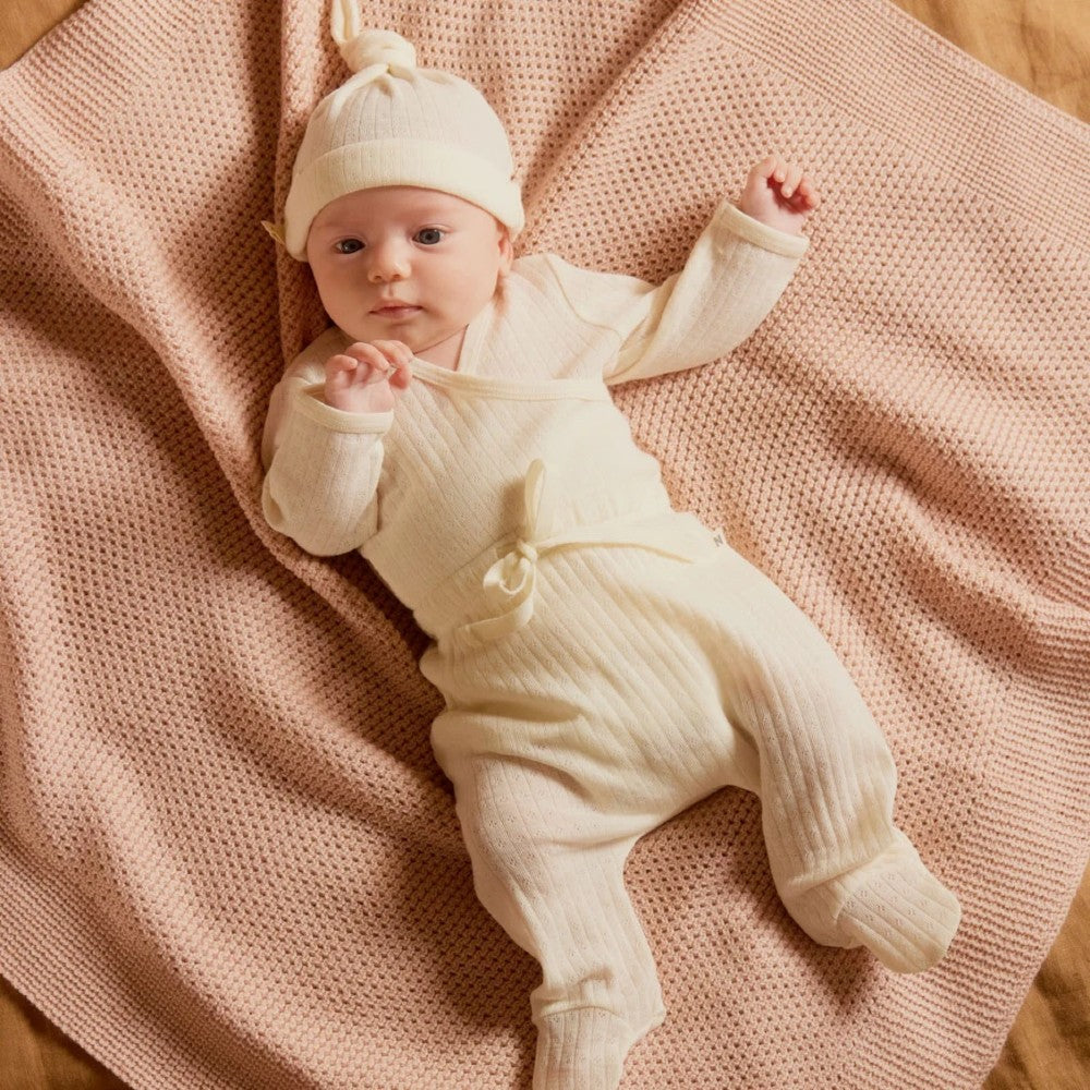 Baby in a cream merino pointelle matching outfit including kimono bodysuit, footed leggings and knotted hat on a pink blanket 