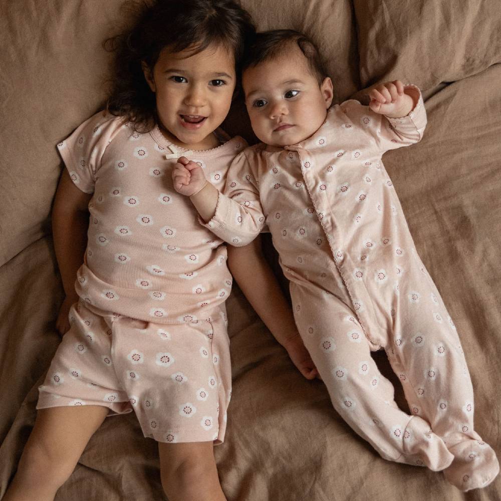Two babies lying side by side on a brown blanket, wearing matching pale pink footed pajamas with a small white floral pattern.