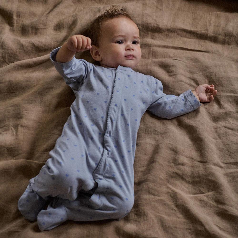 Baby in a light grey footed onesie with blue heart pattern lying on a brown blanket