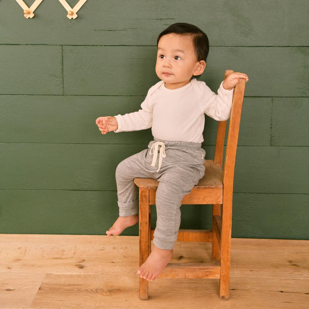 Child sitting on a wooden chair against a green wall wearing a merino cream coloured bodysuit and grey merino leggings