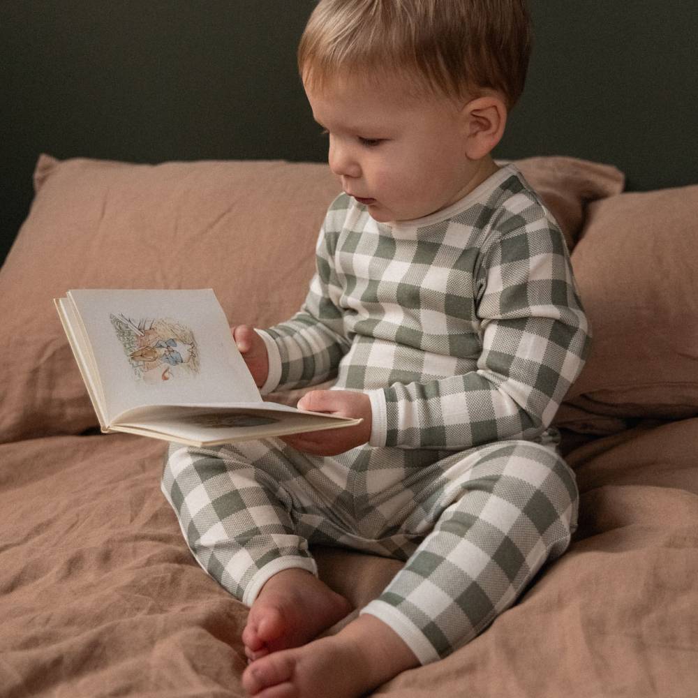 A toddler sitting on a bed reading a book, wearing a green and white gingham check long-sleeve pajama set.