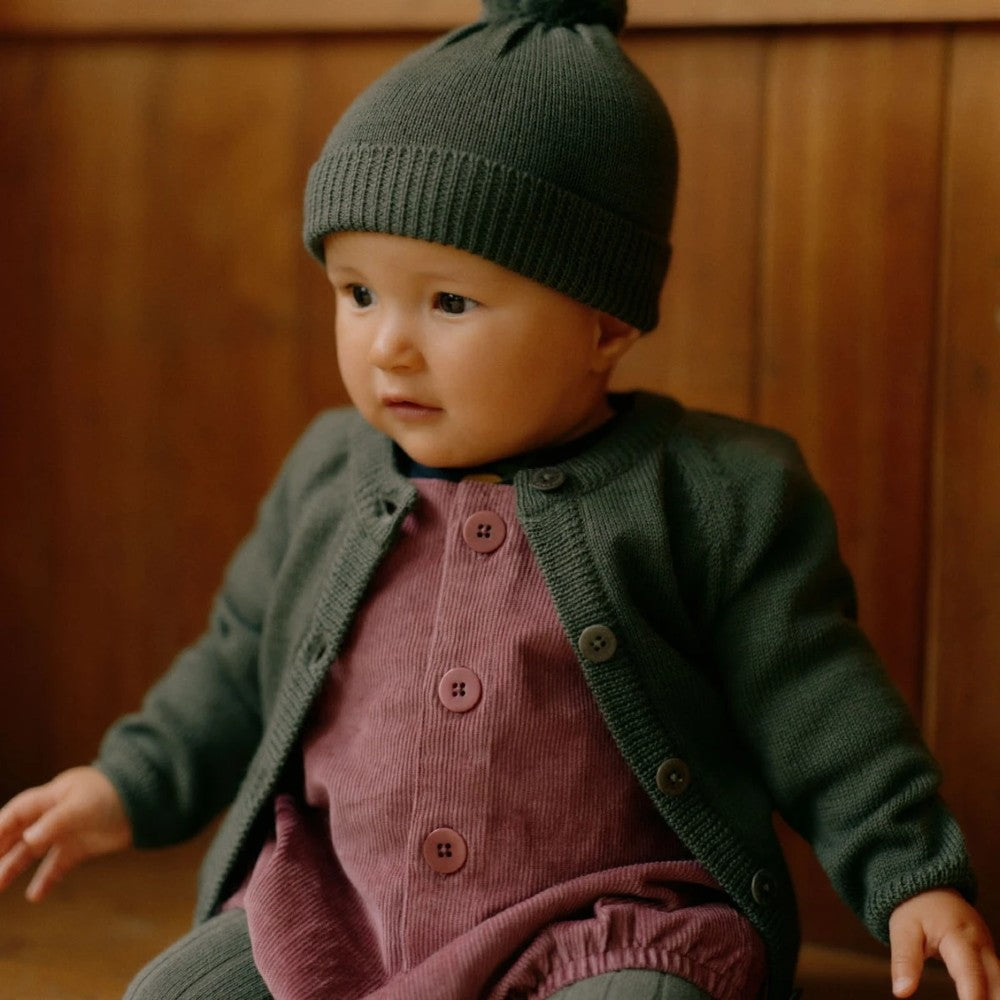 Baby wearing a green merino knitted hat and cardigan sitting against a wooden wall.