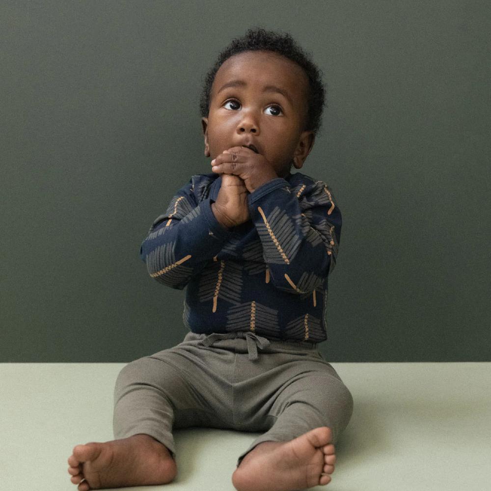 Small child sitting on a light surface with a dark green background and navy bodysuit with tree patterns and green pants