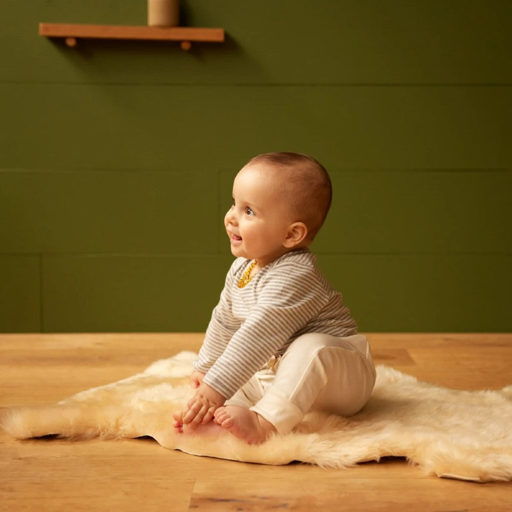 Baby sitting on a lambskin rug wearing a striped bodysuit in a room with a green wall and wooden shelf.