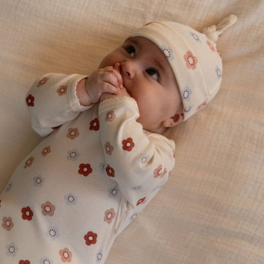 A baby lying on a textured cream blanket, wearing a matching long-sleeve bodysuit and knot beanie with a small floral print in muted red, dusty pink, and light blue.