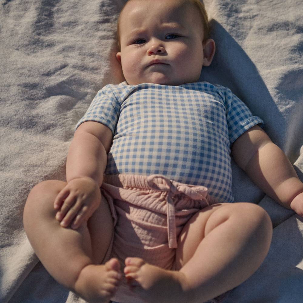 A baby lying on a textured blanket, wearing a light blue and white gingham short-sleeve bodysuit and dusty pink ruffled diaper cover.