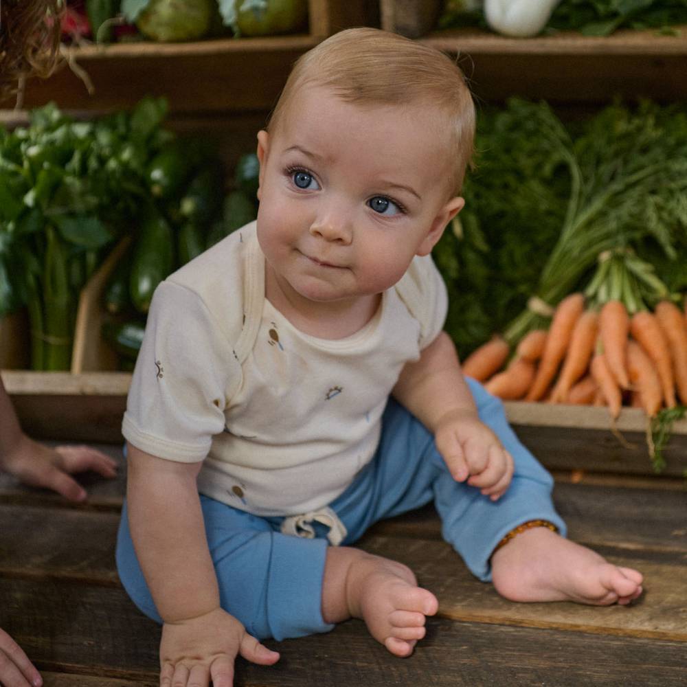 A baby sitting on rustic wooden flooring in a vegetable market setting, wearing a cream short-sleeve bodysuit with light blue pants.