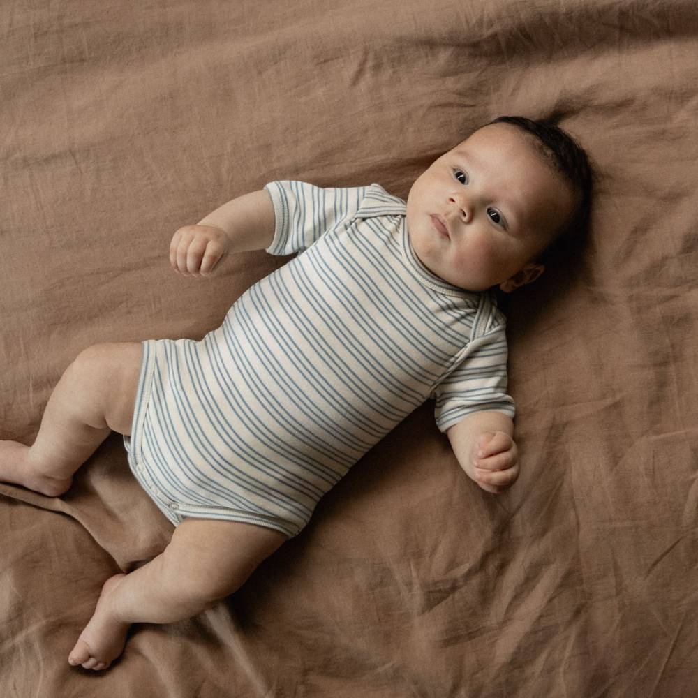 A baby lying on a brown linen blanket, wearing a short-sleeve bodysuit with thin blue and white stripes.