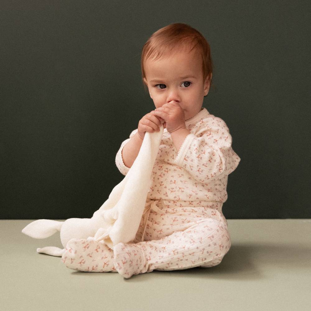 A baby sitting on a light surface against a dark gray background, wearing a cream footed onesie with a small pink floral print and holding a white plush toy.