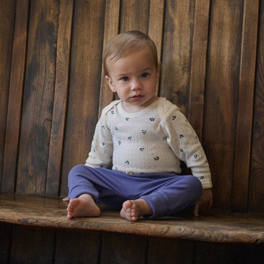 Baby sitting on a wooden bench wearing a white pointelle cotton longsleeve bodysuit with purple berry patterns and purple pants.