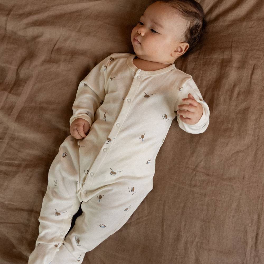 A baby lying on a brown fabric surface wearing a cream-colored long-sleeve footed sleeper with small drums prints.