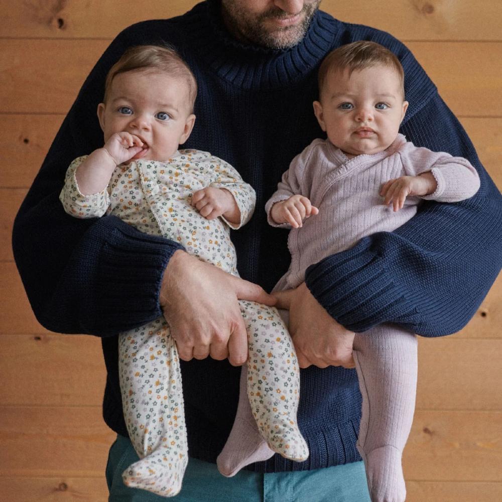 Man holding two babies wearing a floral onesie and purple stretch and grow against a wooden background