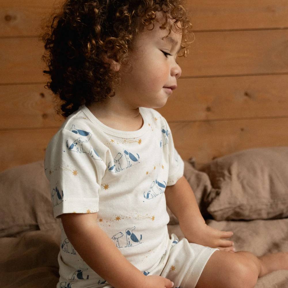 A toddler with curly brown hair sits on a bed wearing a cream short-sleeve pajama top and matching shorts with a blue and gold dog print, set against a warm wood-paneled background.