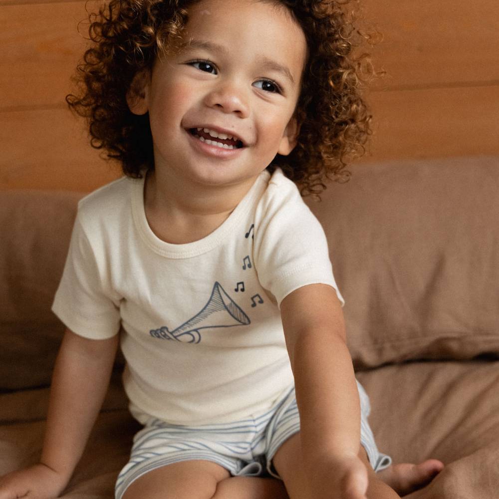 A smiling toddler with curly brown hair sits on a brown couch wearing a cream short-sleeve t-shirt with a blue trumpet graphic and striped shorts.