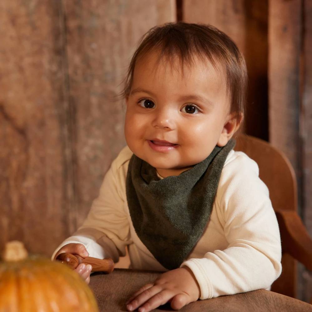 Child wearing a green cotton terry bandana bib sitting at a table with a pumpkin, smiling.