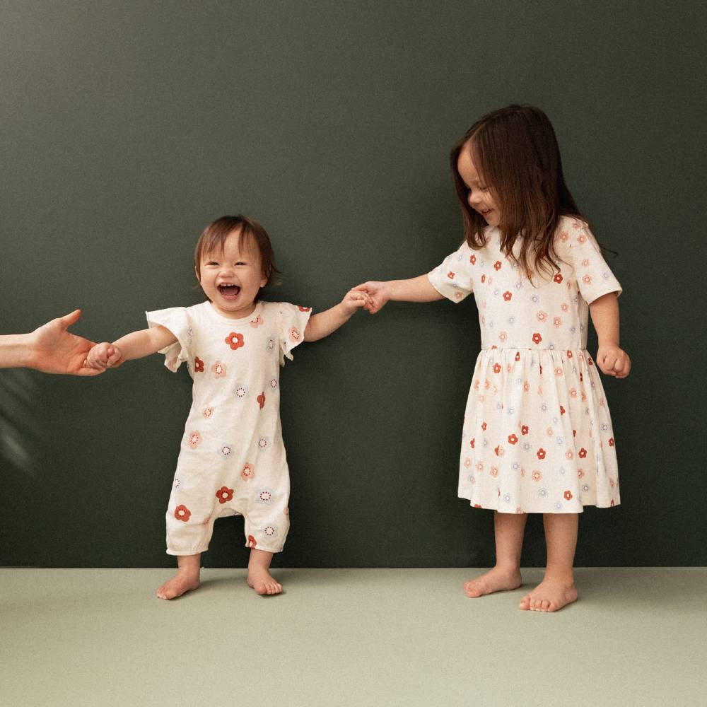 Two young children standing barefoot and holding hands against a dark green wall. The child on the left wears a cream short-sleeve onesie with small orange and blue floral prints, and the child on the right wears a matching cream dress with short sleeves and a gathered waist.