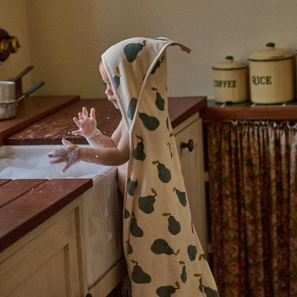 Child in a kitchen wearing a cream cotton hooded towel with pear pattern, standing by a sink.