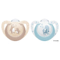 Two NUK baby pacifiers on a white background, one in beige with a penguin design and one in light blue with a dinosaur design. Small text on the right reads '0-6 months'.