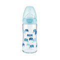 A NUK baby bottle with a clear glass body decorated with blue and light blue elephant patterns and the name 'NUK' printed in blue. It features a light blue collar and a translucent light blue silicone nipple on a solid white background.