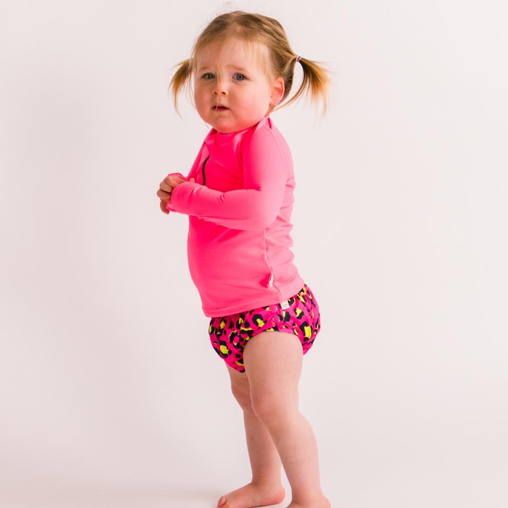 Child wearing a neon pink long-sleeve rash vest and colorful swim nappy on a plain background