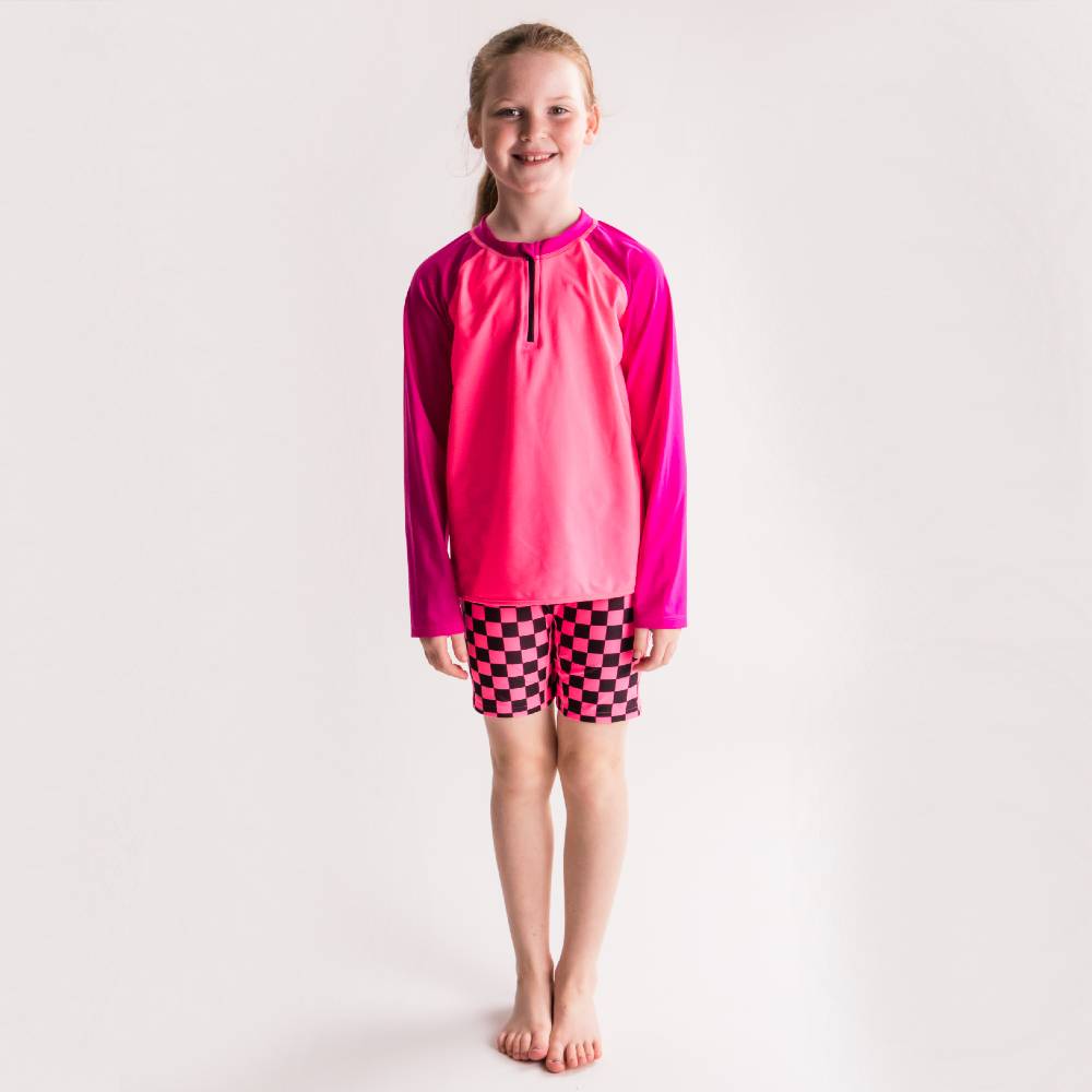 Young girl wearing a bright pink long-sleeve rash vest and checkered swim shorts on a white background