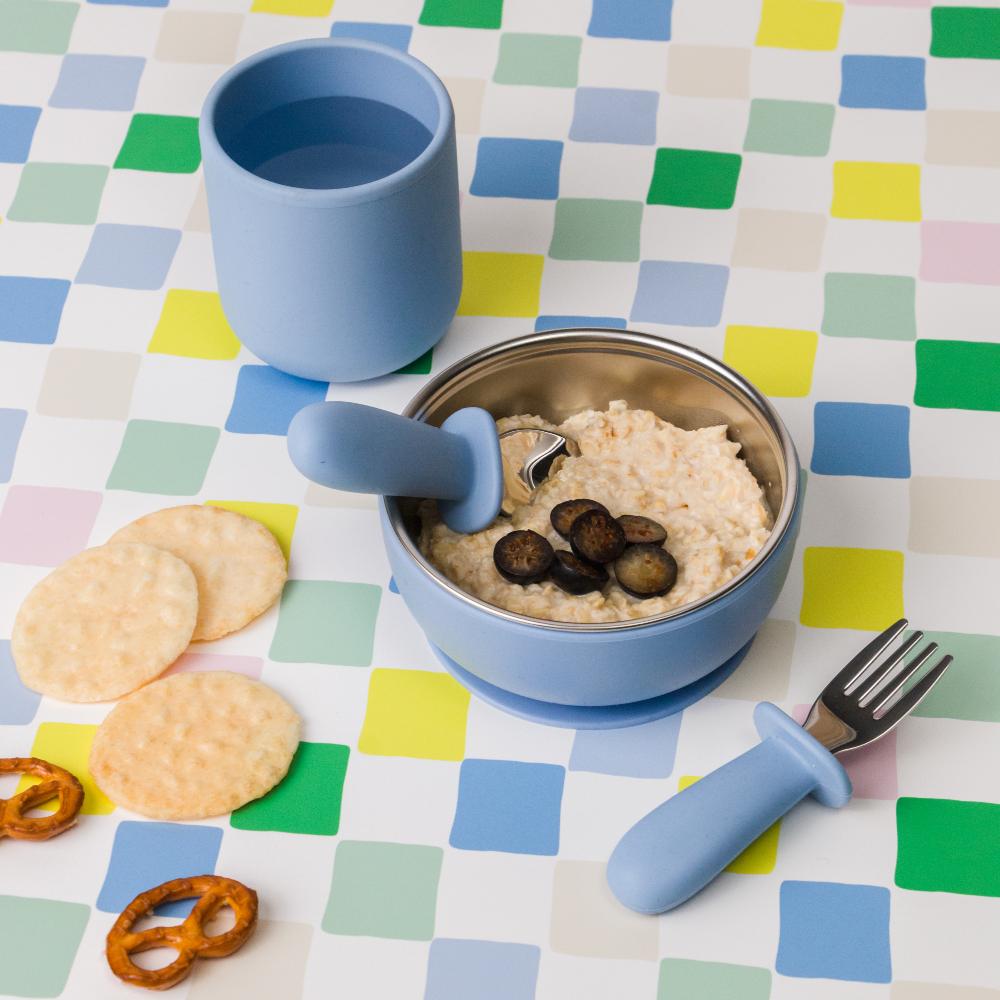 Children's tableware set with blue silicone cup, bowl, and utensils on a checkered tablecloth.