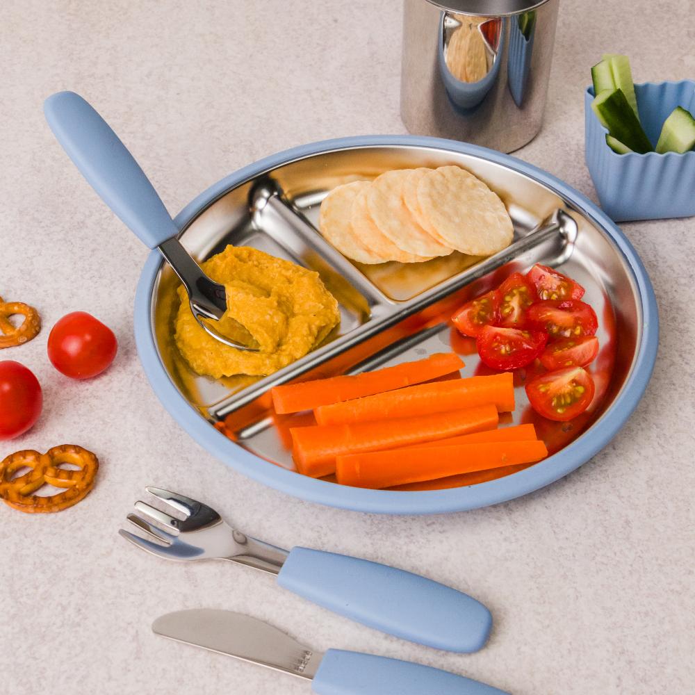 Children's stainless steel plate with blue handles, cutlery, and various food items on a light surface.