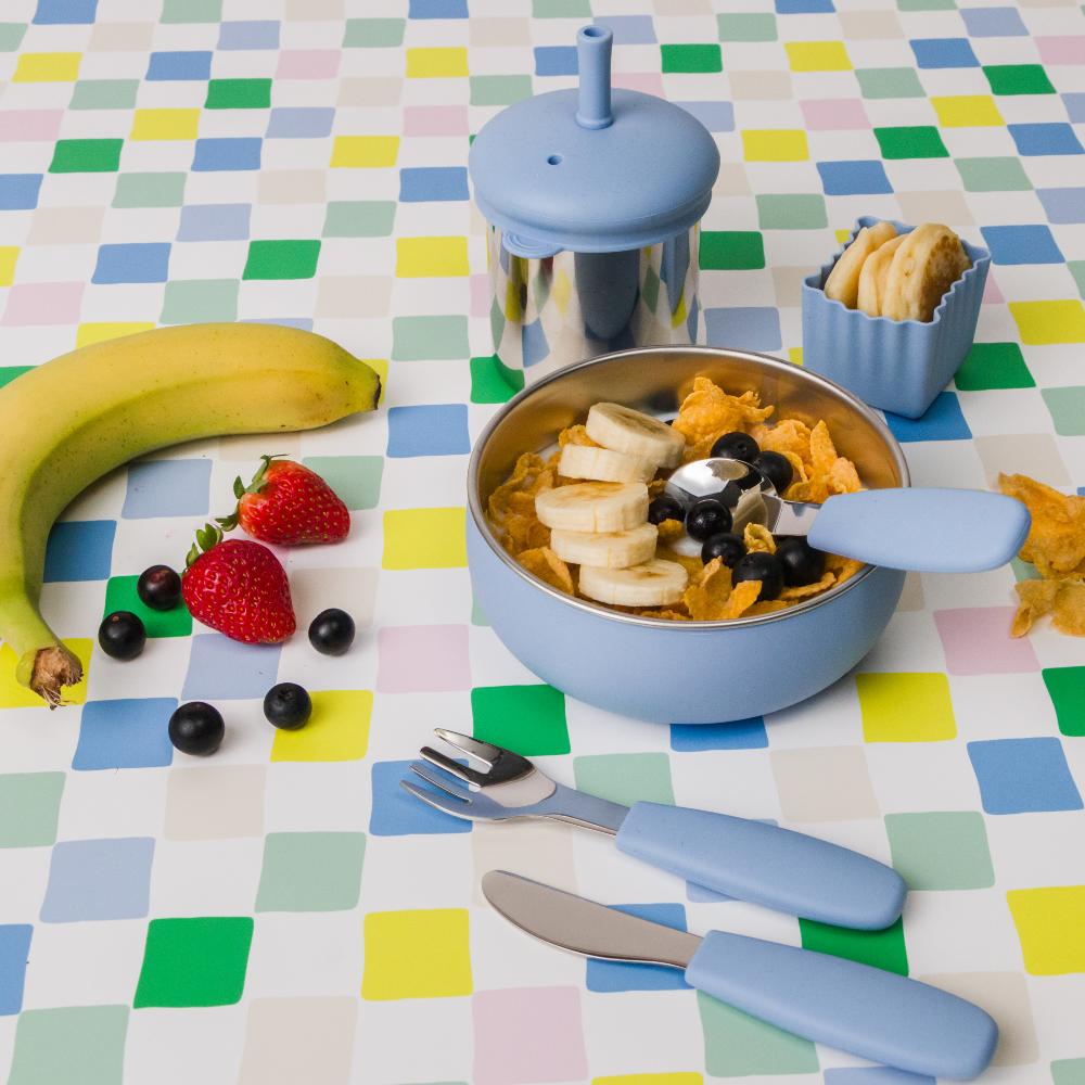 Nestling baby feeding set with a stainless steel bowl of cereal, fruit, and cutlery on a checkered tablecloth.