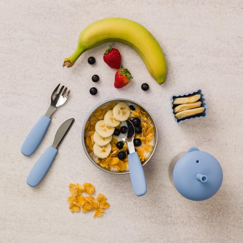 Breakfast setup with stainless steel bowl, bananas, strawberries, and utensils on a beige surface