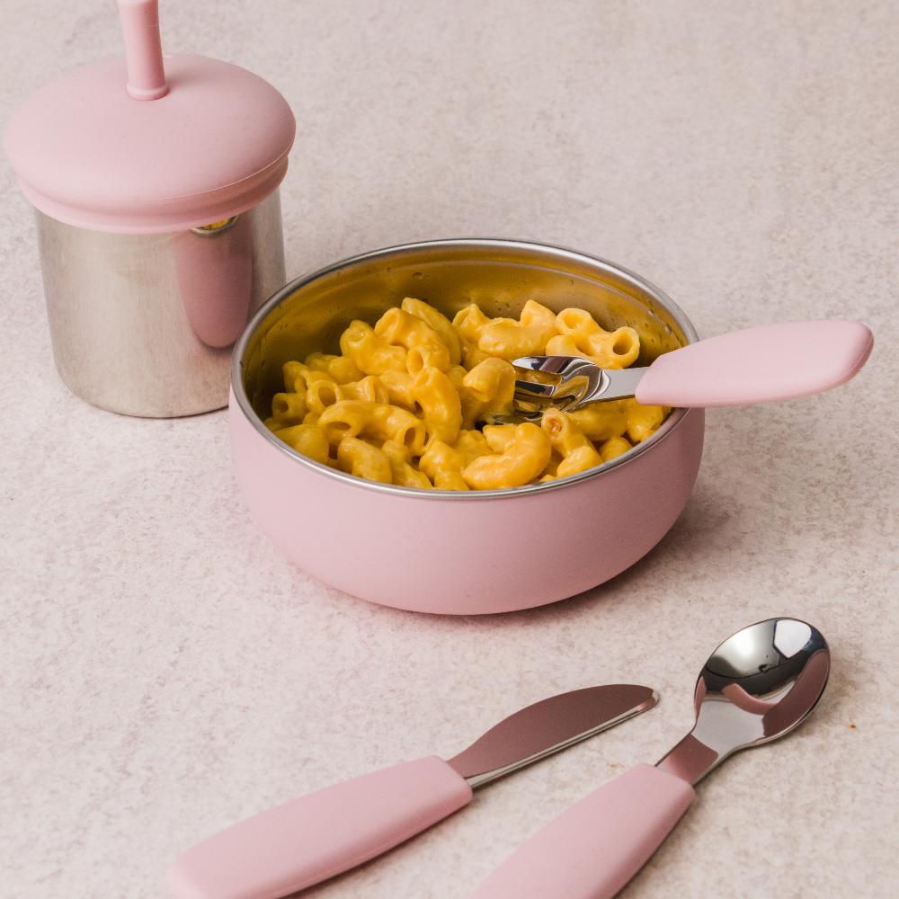 Pink silicone and stainless steel bowl with macaroni and cheese, spoon, and fork on a beige surface