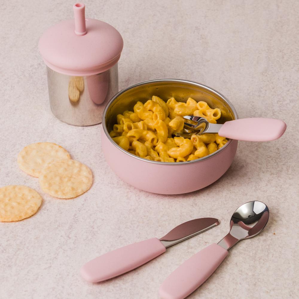 Pink stainless steel and silicone baby bowl with macaroni and cheese, pink cup, and cutlery on a beige surface.