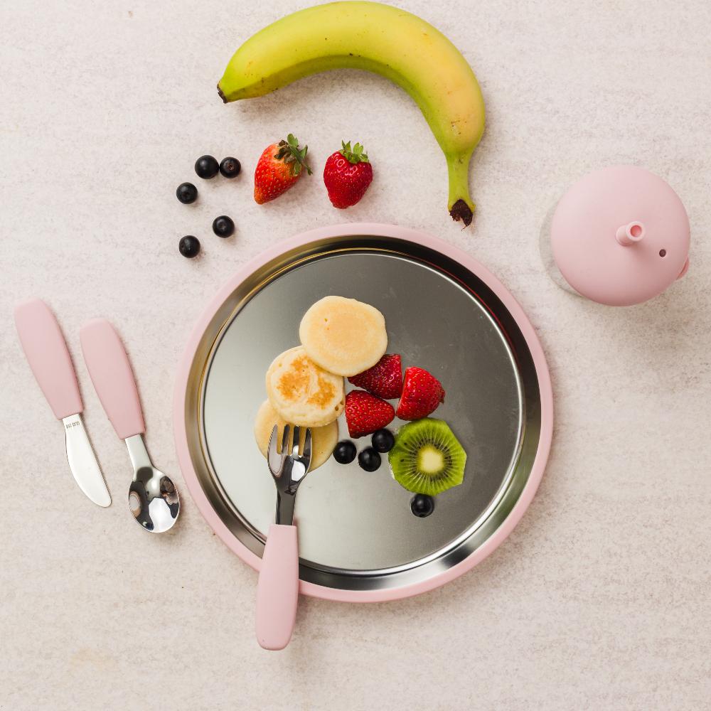 Fruit and pancake stack on a pink stainless steel plate with utensils and a banana.