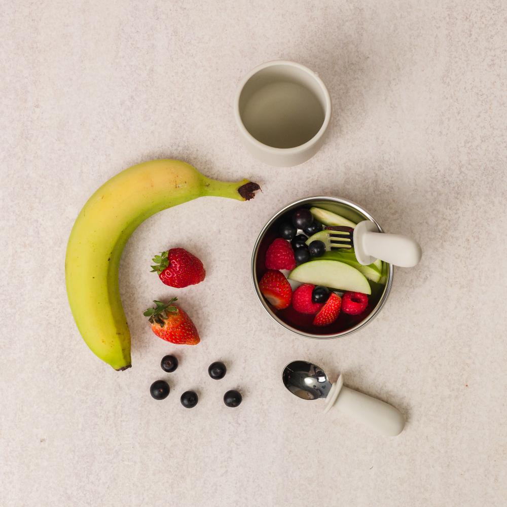 Fruits and a bowl of fruit salad on a textured surface with nestling cup and stainless steel cutlery