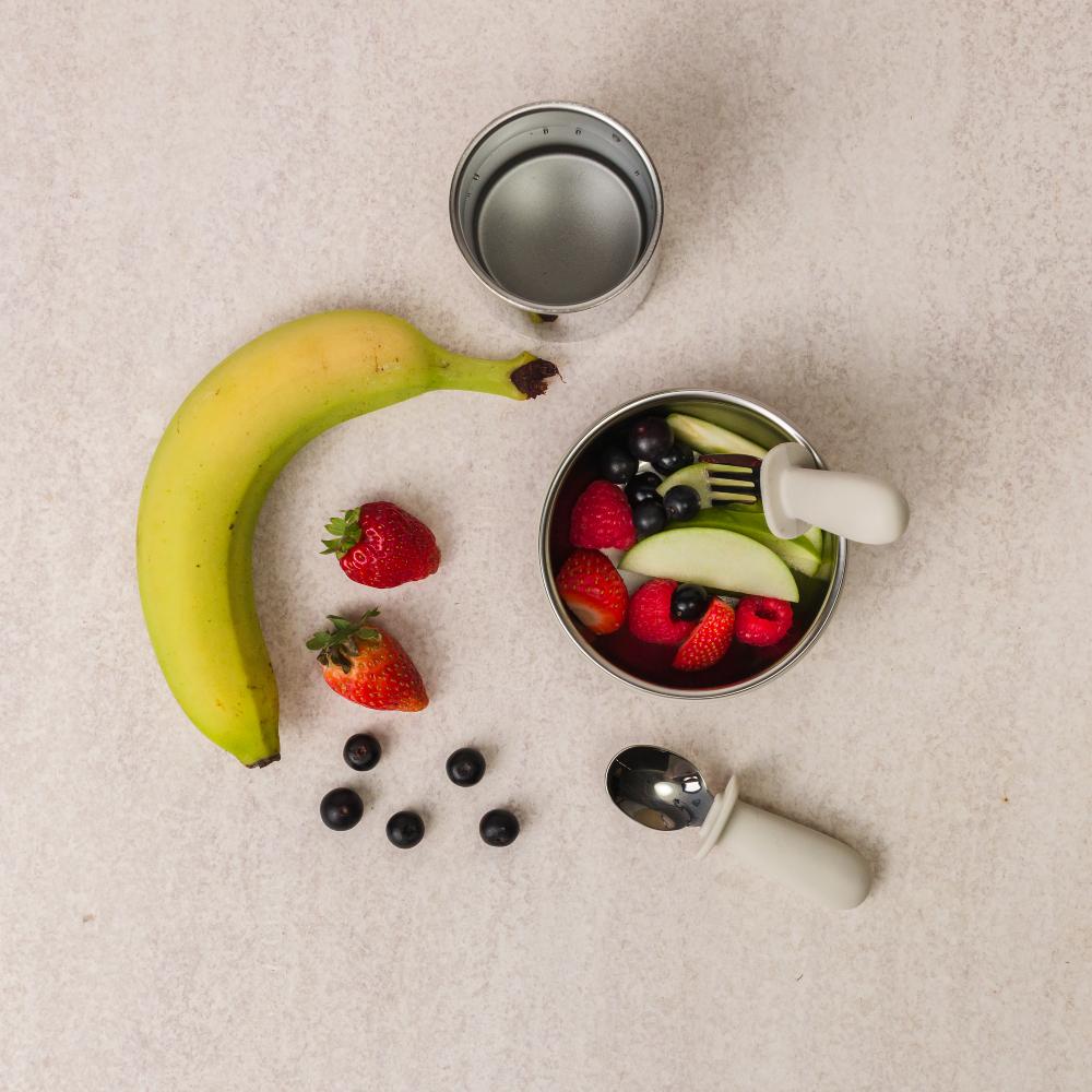 Fruit salad with a baby spoon, fork, stainless steel cup, banana, and berries on a beige surface