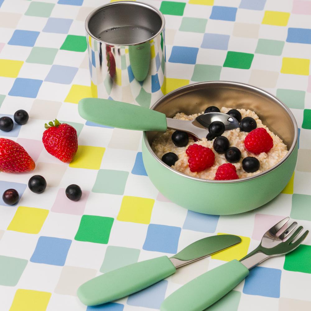 Toddler stainless steel green bowl and cutlery with food, utensils, and berries on a colorful checkered tablecloth