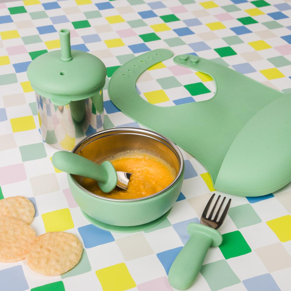 Green  Nestling baby tableware set with bowl, bib, cup and cutlery on a colorful checkered tablecloth