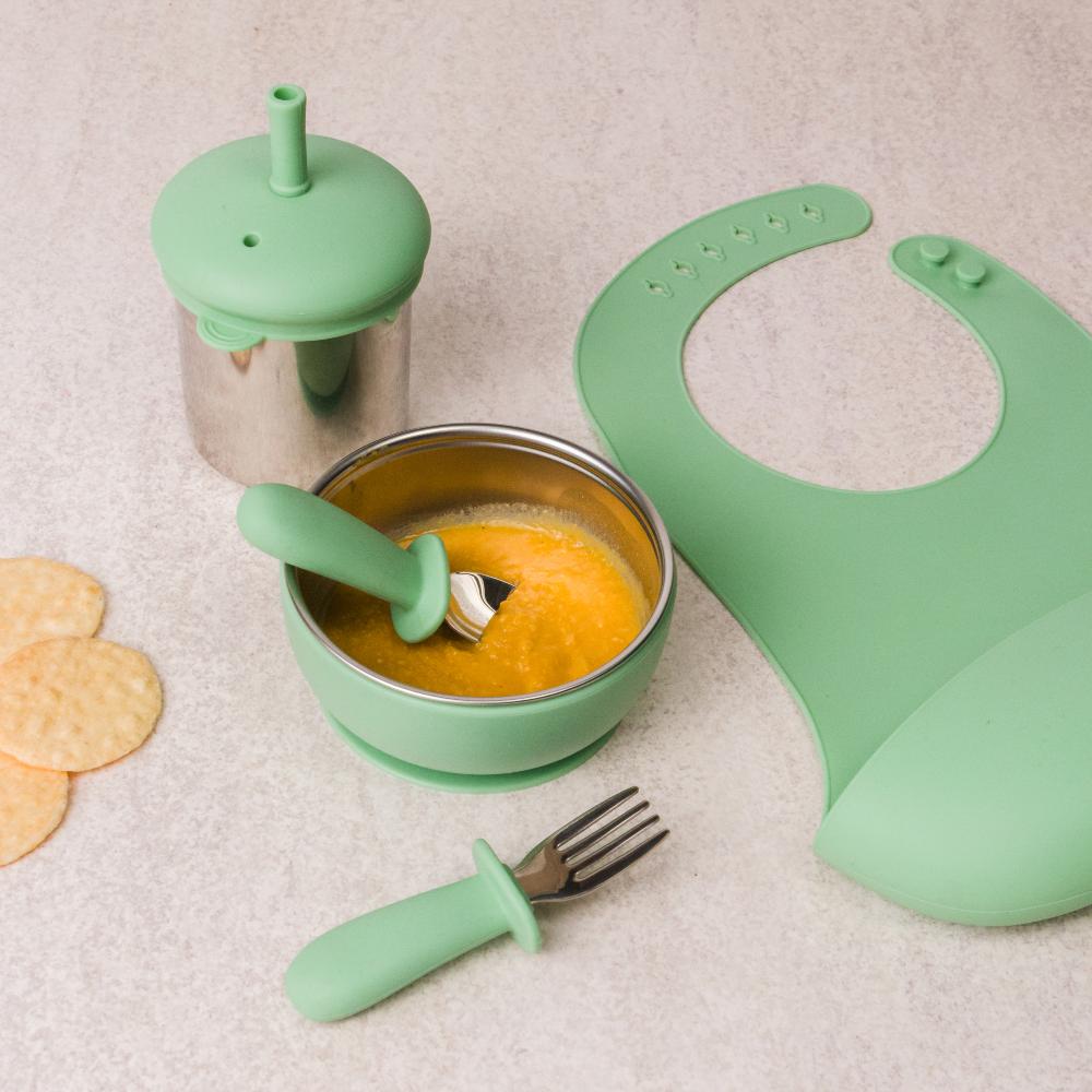 Sage green stainless steel baby feeding set with bowl, spoon, bib, and fork on a neutral background