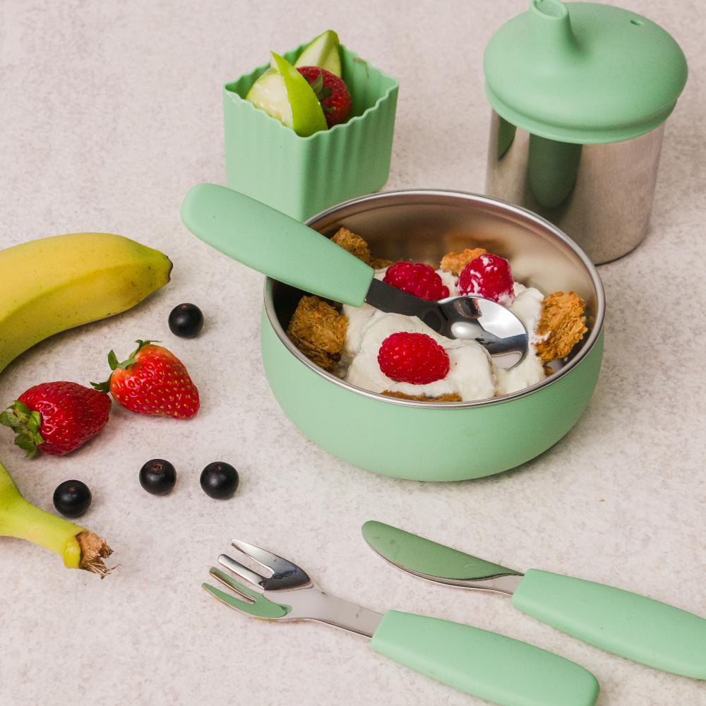 Sage green Nestling baby feeding set with bowl container, fork, knife, and spoon on a light surface with fruits.