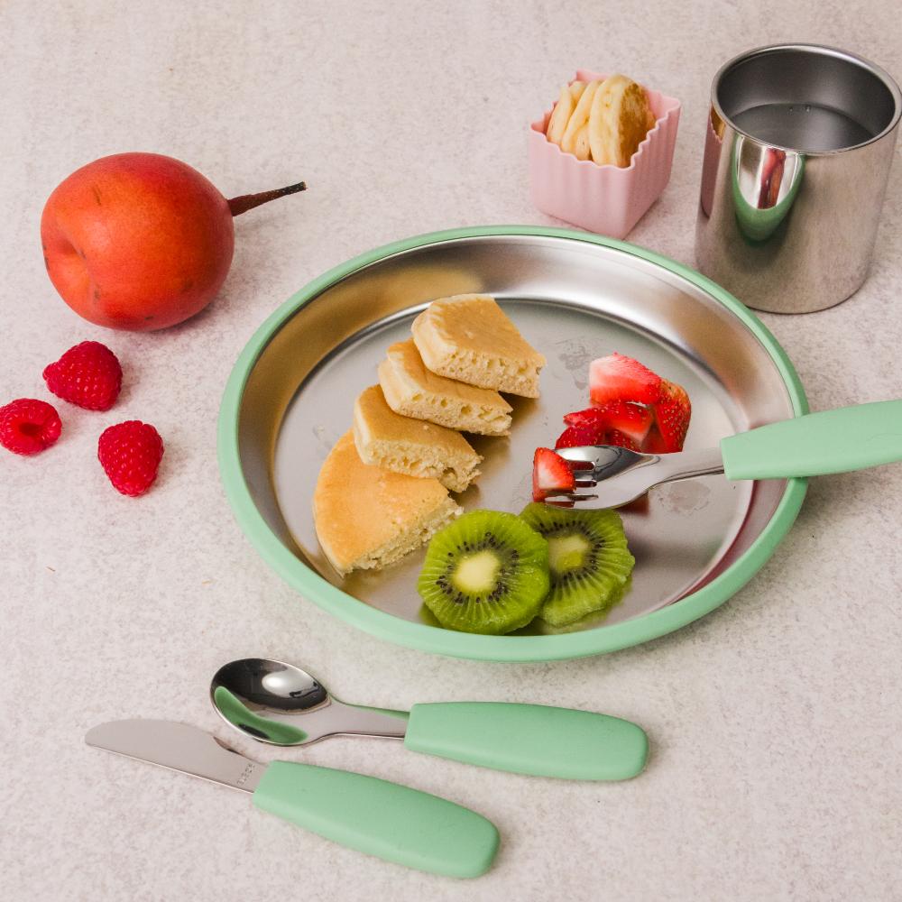 Children's sage green stainless steel plate with food, cutlery, and fruits on a light surface