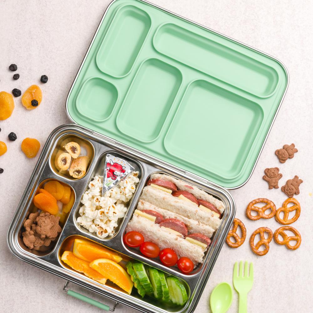 Green bento box with compartments filled with food on a light surface with snacks around.