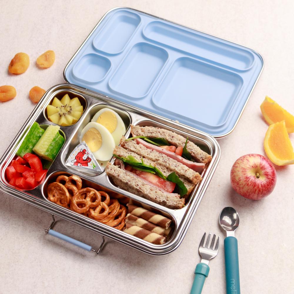 Lunchbox with compartments filled with food, including sandwiches and fruits, on a light surface.