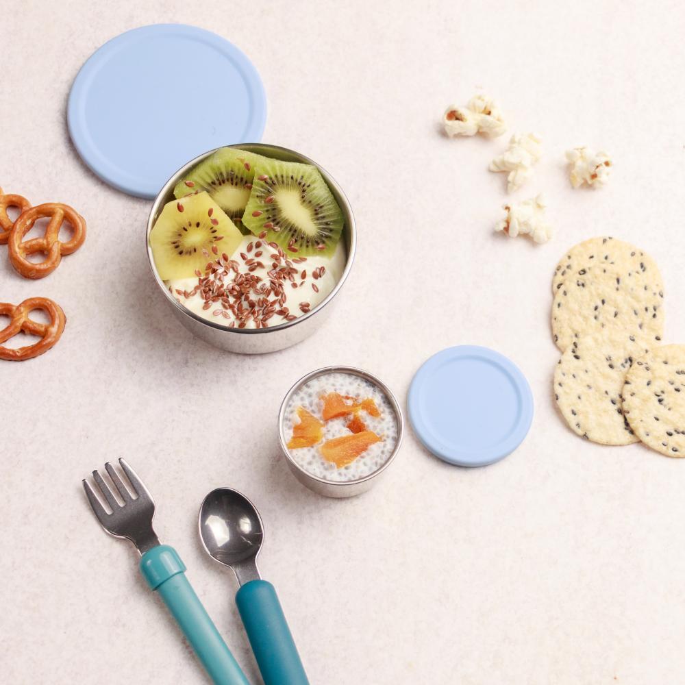 Two small containers with lids, a bowl of fruit, and snacks on a light surface.