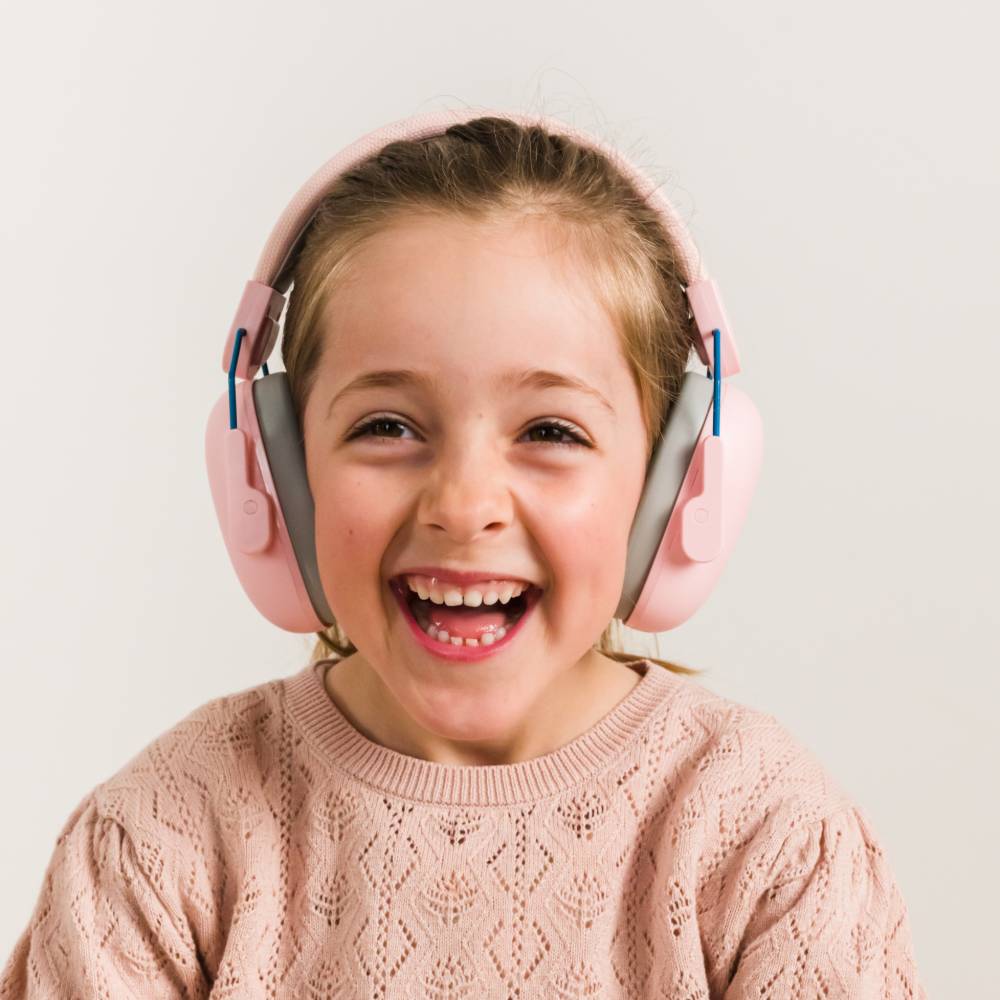 A young girl with blonde hair tied back is smiling broadly while wearing pink over-ear muffs with gray inner padding and small blue accents. 
