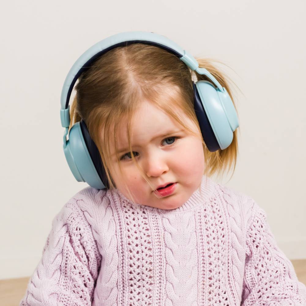 A young child wearing light blue ear muffs and a pale pink cable-knit sweater, photographed against a plain light beige wall.