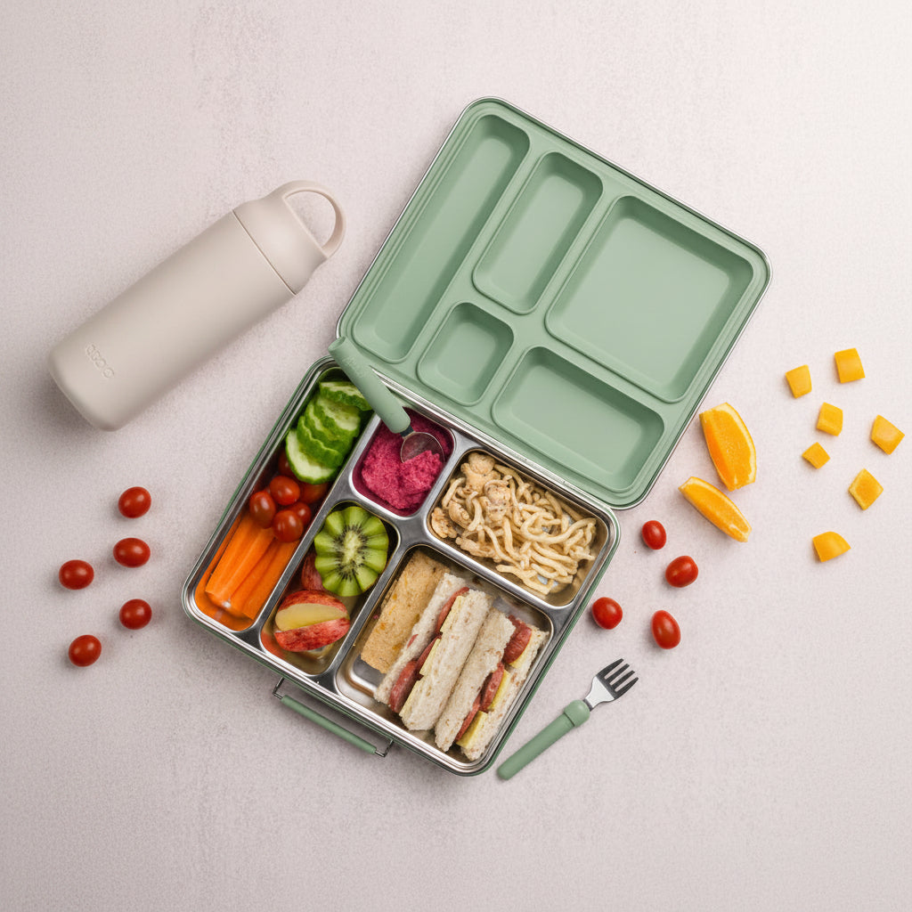 A light green compartment bento lunch box with a stainless steel tray, filled with a sandwich, pasta, sliced fruit, carrots, and a small dip, arranged on a light beige surface with a bottle, fork, and orange slices nearby.